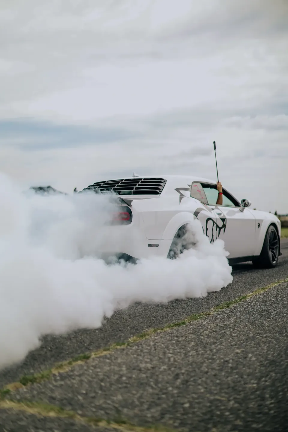 Bílý Dodge Challenger zezadu v oblaku kouře, řidič zvedá ruku z okna