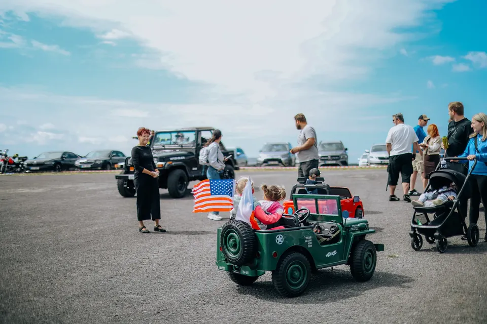 Děti v zeleném elektrickém Jeepu s americkou vlajkou, kolem návštěvníci festivalu