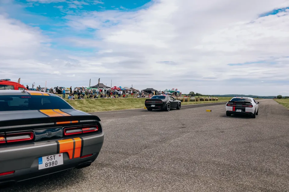 Dva Dodge Challengery jedou po dráze, v pozadí diváci