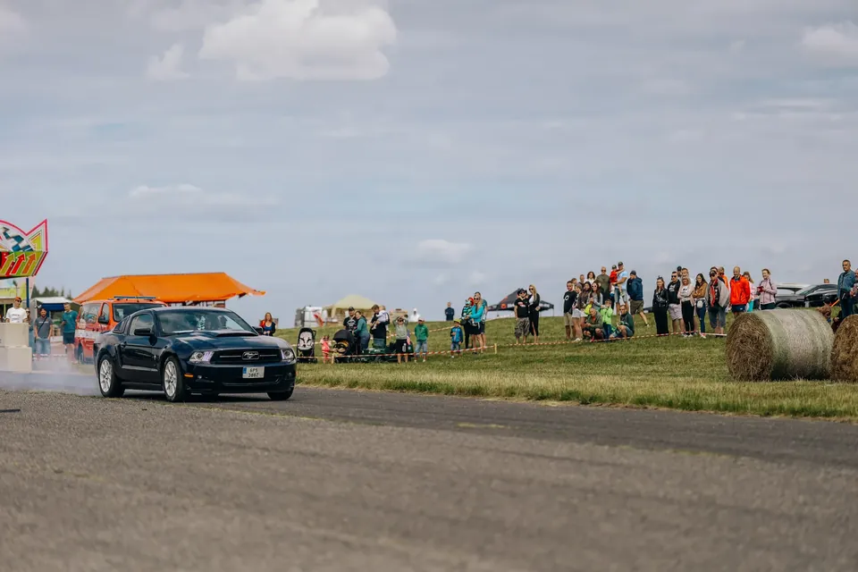 Černý Ford Mustang jede po dráze, v pozadí diváci a oranžový stan