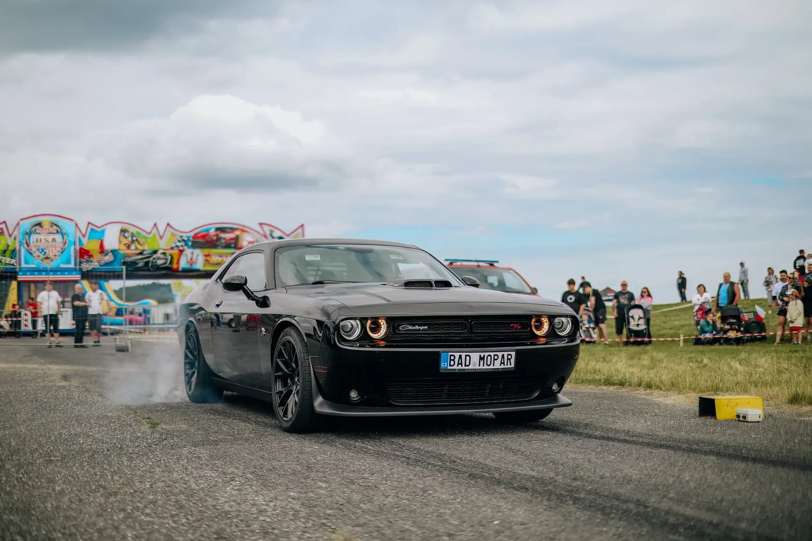 Černý Dodge Challenger vyjíždí z kouře na dráze, v pozadí diváci a pouťová atrakce